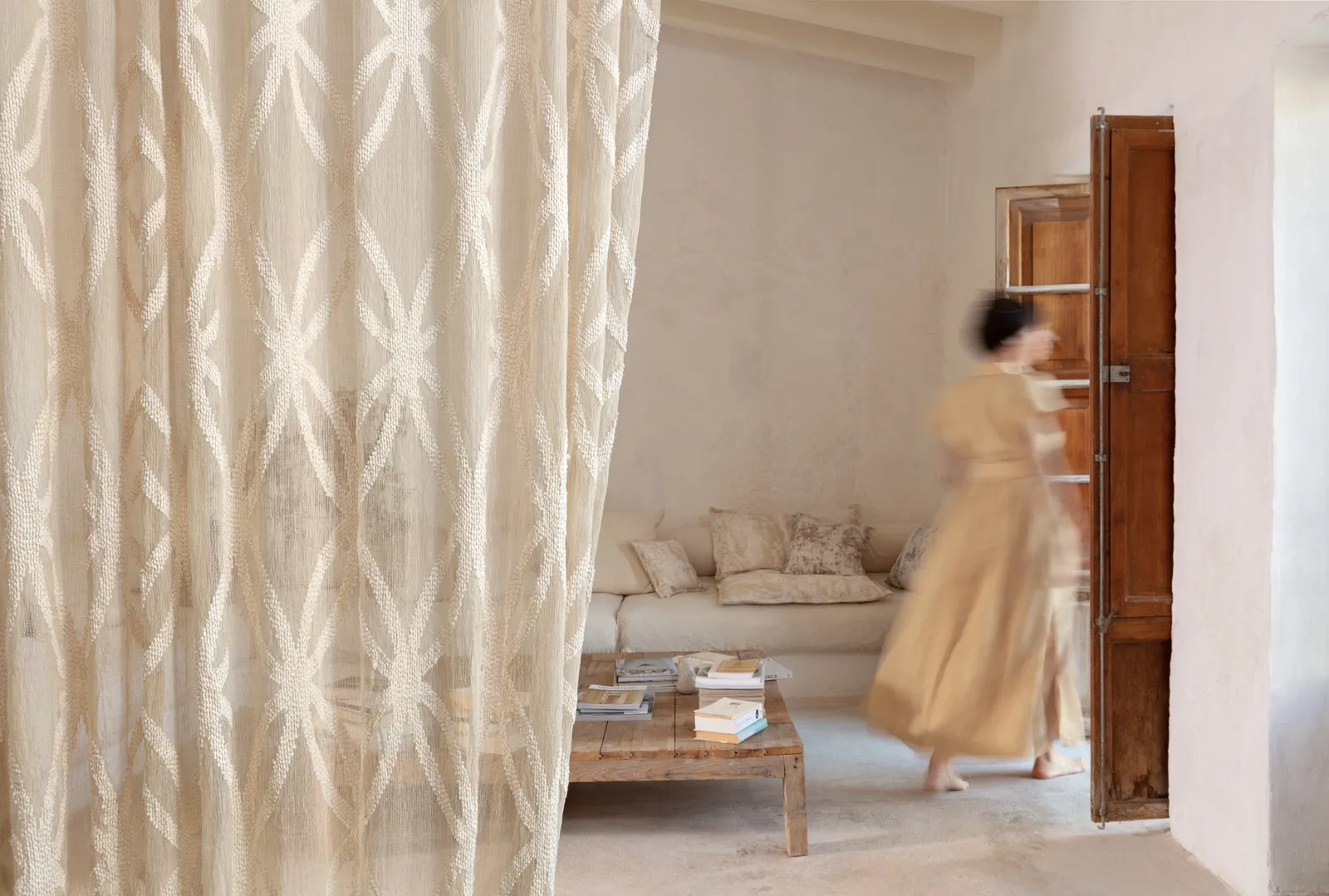 Woman walking through a sunlit living room behind a sheer patterned curtain